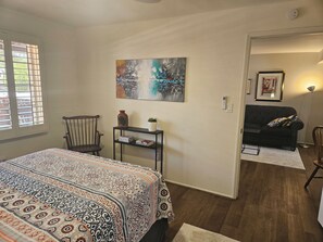 A bedroom with a bed, a chair, a small table with a vase, a painting, and a sofa in the adjacent room.