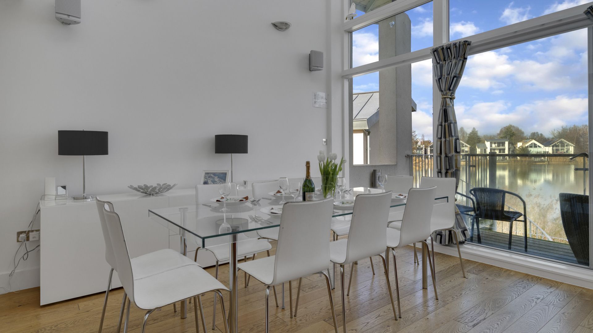 A modern dining area with a glass table and white chairs, large windows with a view of a lake and houses, and a flat-screen TV mounted on the wall.