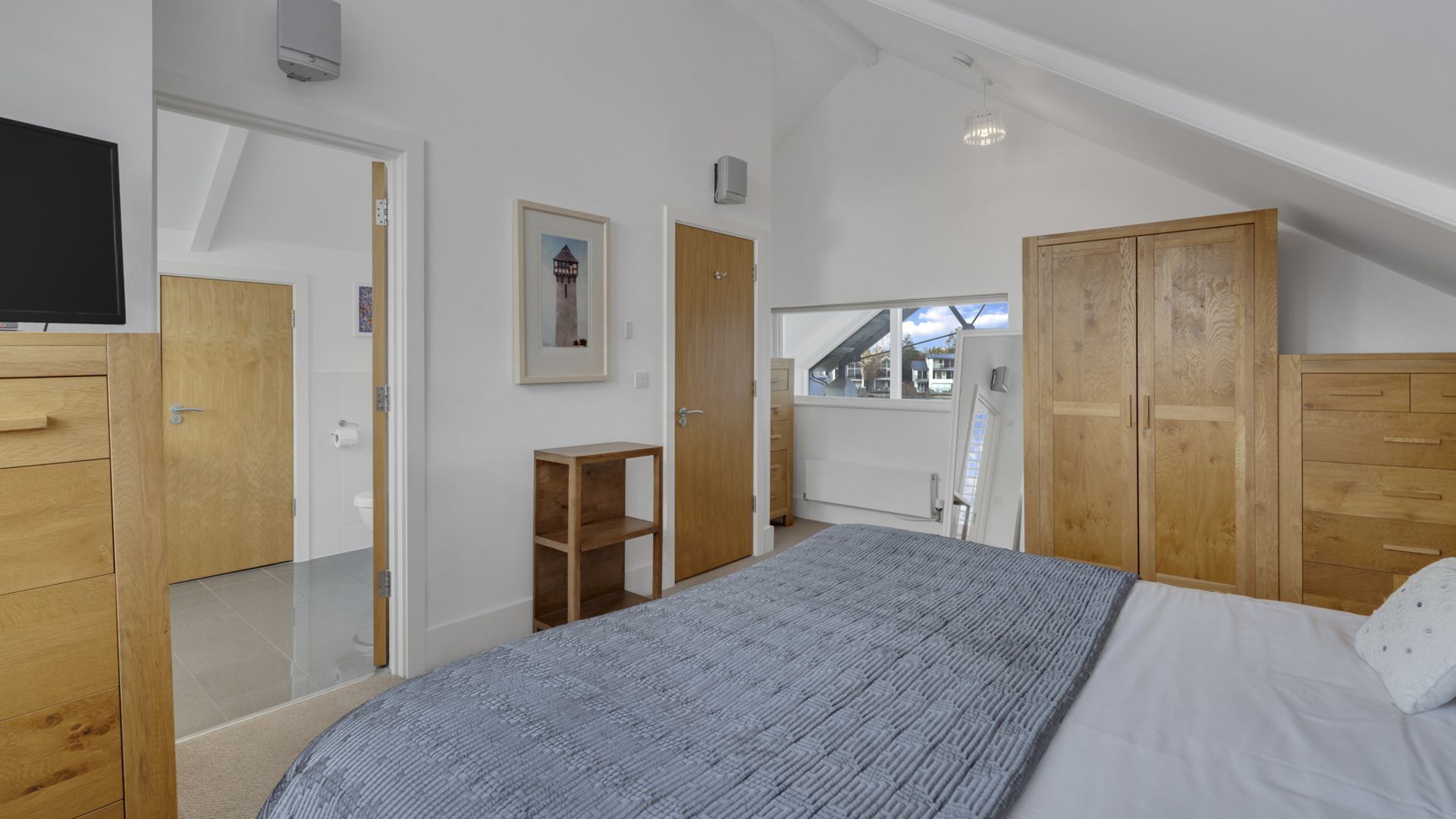 A bedroom with a bed, wooden wardrobe, a small desk, and a window with a view of houses.