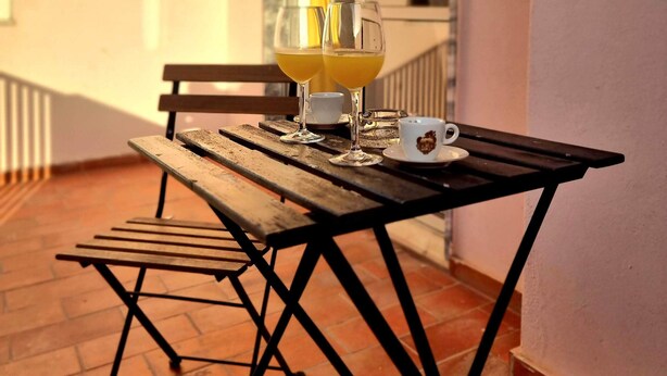 A wooden table with two glasses of orange juice, a white cup, and a saucer on a balcony with a tiled floor and a bench. - Happy House Central Apartment (Faro)