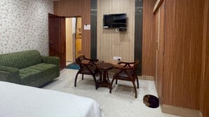 A hotel room with a bed, a green sofa, a wooden dining table with chairs, a TV mounted on the wall, and a wardrobe.