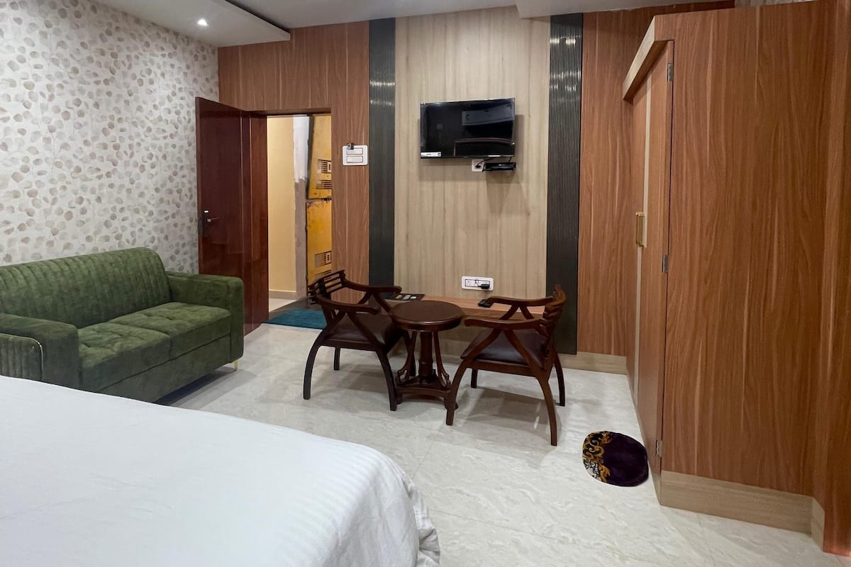 A hotel room with a bed, a green sofa, a wooden dining table with chairs, a TV mounted on the wall, and a wardrobe.