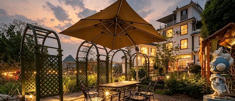 Un patio exterior con una mesa y sillas, un paraguas grande y vista a un edificio al fondo.