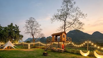 A wooden observation tower with a staircase, surrounded by trees and a tent, set against a mountainous backdrop.
