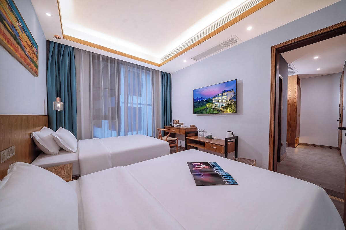 Habitación de hotel con dos camas, un escritorio, una silla, un cuadro y vista a un edificio por la ventana.