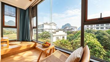 A modern room with a large window, a wooden table with a fruit bowl, a woven chair, and a view of traditional buildings and greenery.