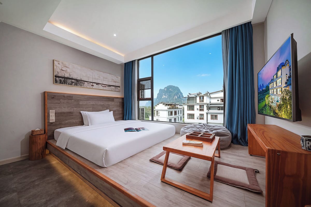 Habitación de hotel moderna con una cama grande, un escritorio de madera, televisión y vistas a edificios a través de amplios ventanales.