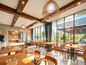 A spacious dining area with wooden tables and chairs, large windows offering a view of greenery, and a ceiling with exposed beams and woven light fixtures. - Yangshuo Floral Whisper Valley (Guilin)