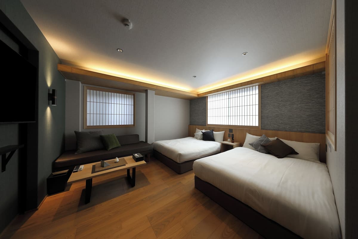 A modern hotel room with two beds, a sofa, a desk, and large windows.
