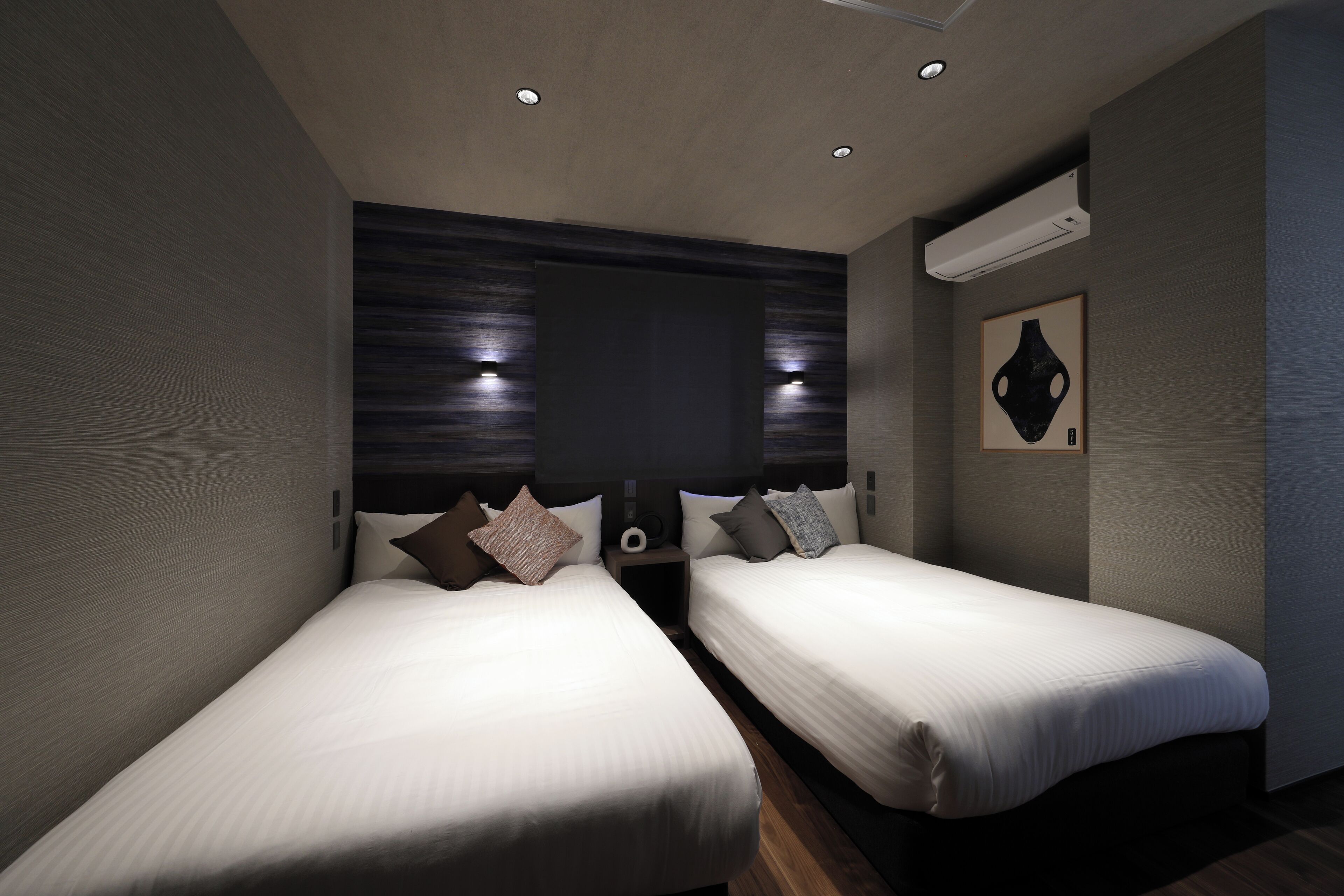 A modern hotel room with two beds, a dark wood headboard, a wall-mounted air conditioner, and a framed artwork.