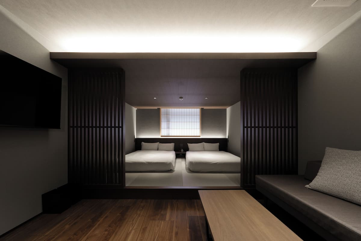 A compact room with two beds, a sofa, a TV, and a window with blinds.