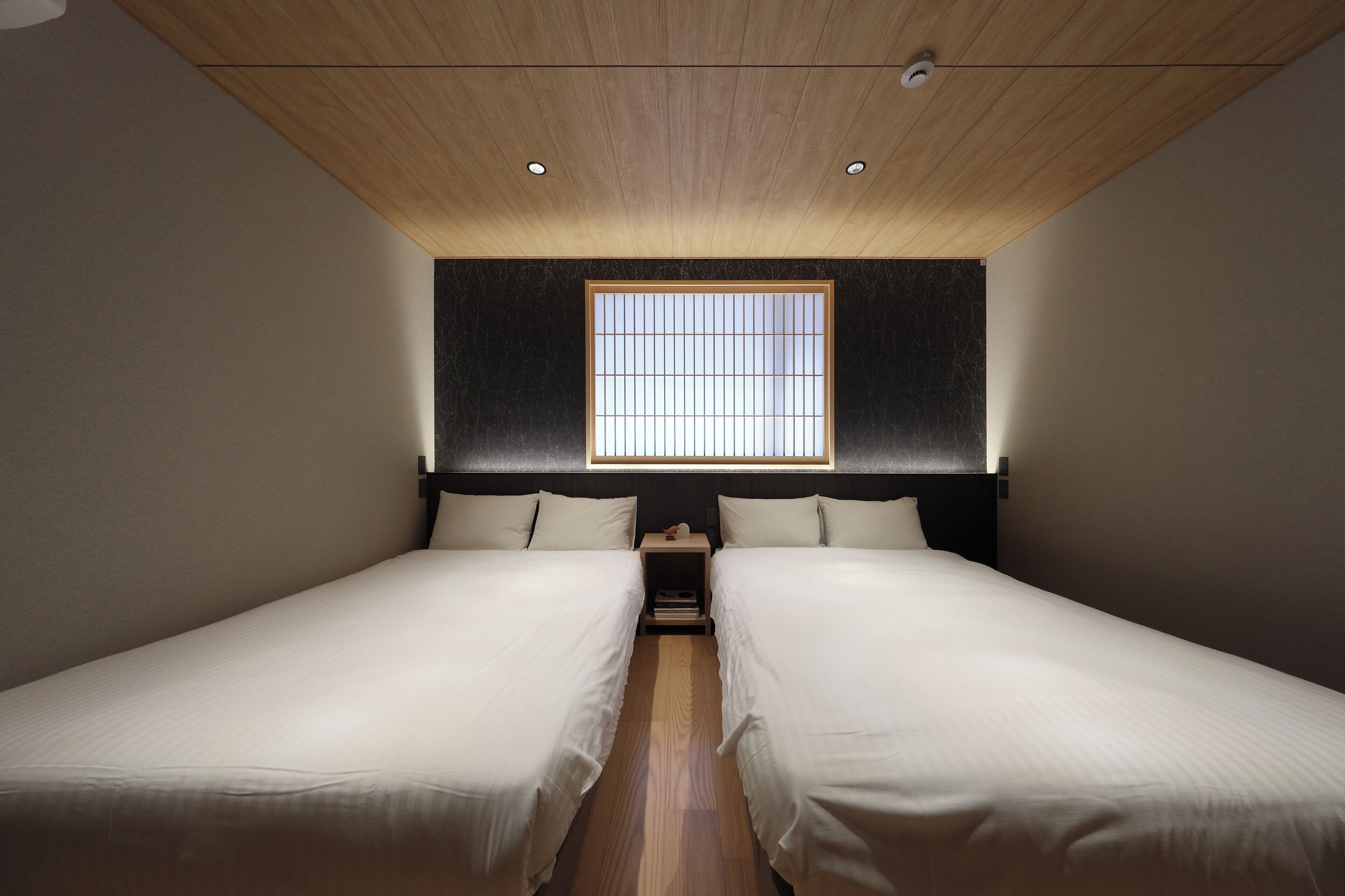 A room with two beds, a wooden ceiling, a window with vertical blinds, and a nightstand between the beds.