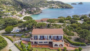 En ejendom ved kysten med et stort hus, betontag, terrasse med udendørs møbler og udsigt over havet og de omkringliggende bakker.