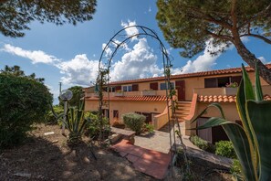 Un edificio de dos pisos con tejado de tejas rojas, un camino de piedra que conduce a la entrada y un jardín con diversas plantas y un arco de metal.