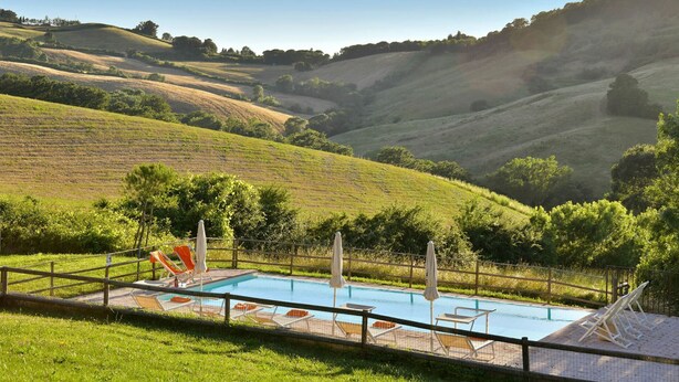 Una piscina con sillones de descanso y sombrillas, rodeada por una valla, con colinas y árboles en el fondo.