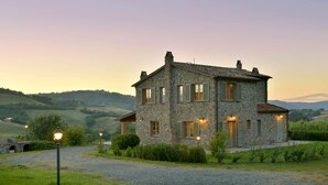 Una casa de piedra con camino de grava, rodeada de vegetación y colinas al fondo.