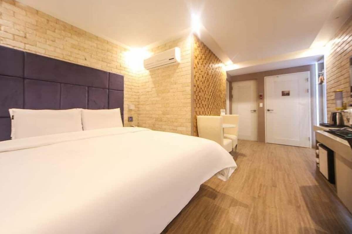 A modern hotel room with a large bed, a brick accent wall, a wooden floor, a small seating area, and a kitchenette.