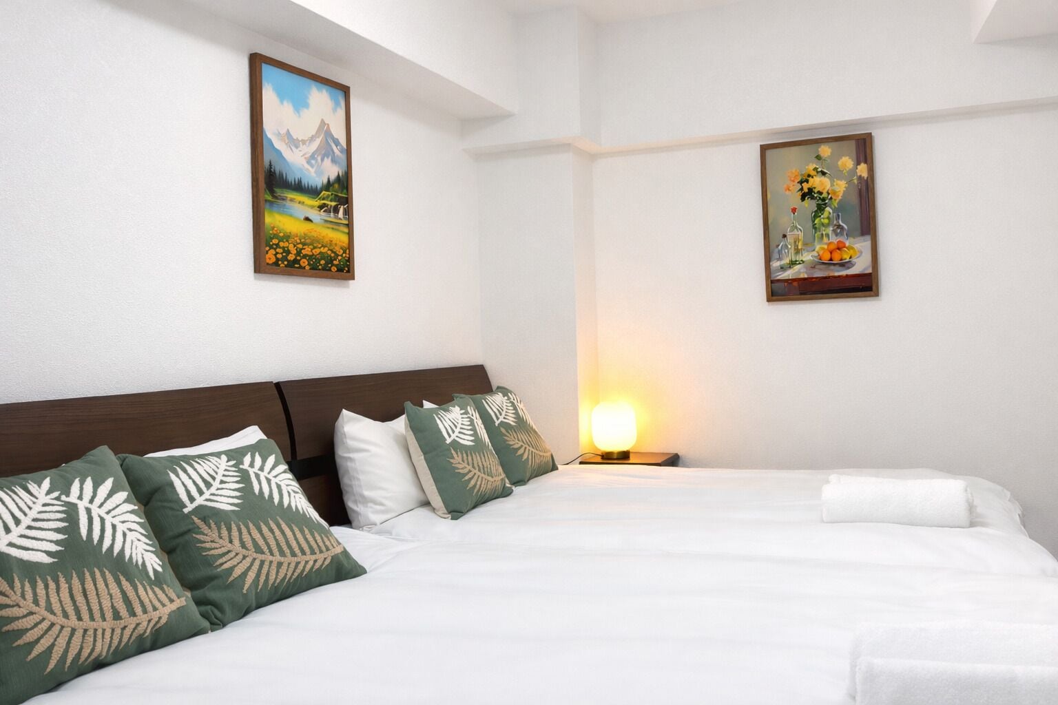A hotel room with a large bed, two pillows, a nightstand with a lamp, and two framed pictures on the wall.
