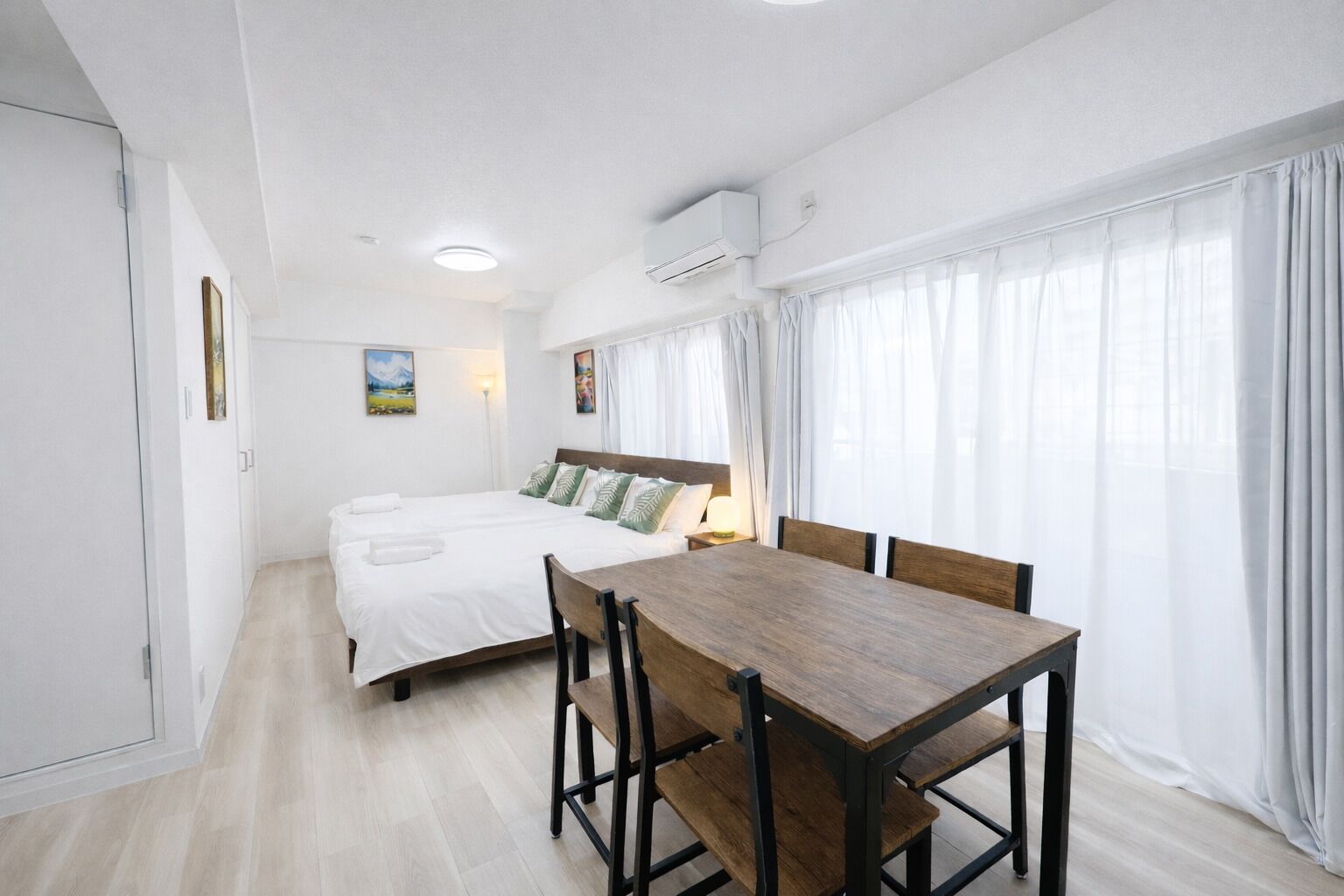 A hotel room with a bed, a wooden dining table, and a window with sheer curtains.