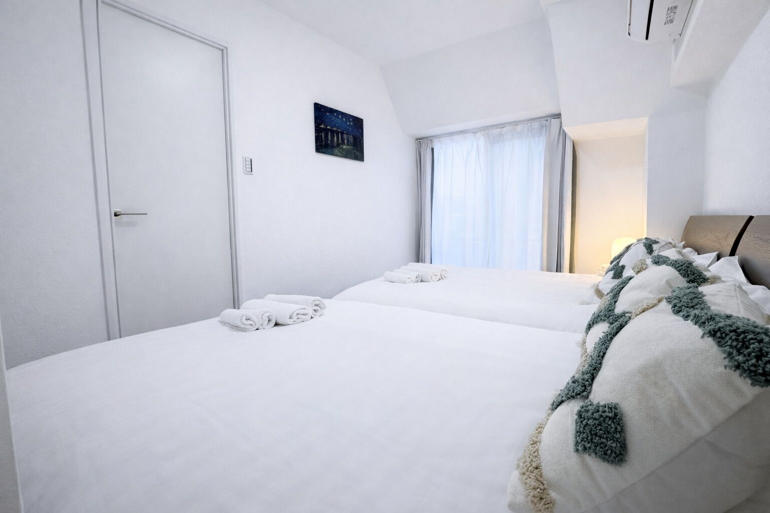 A hotel room with two beds, white linens, a night sky painting, and a window with sheer curtains.