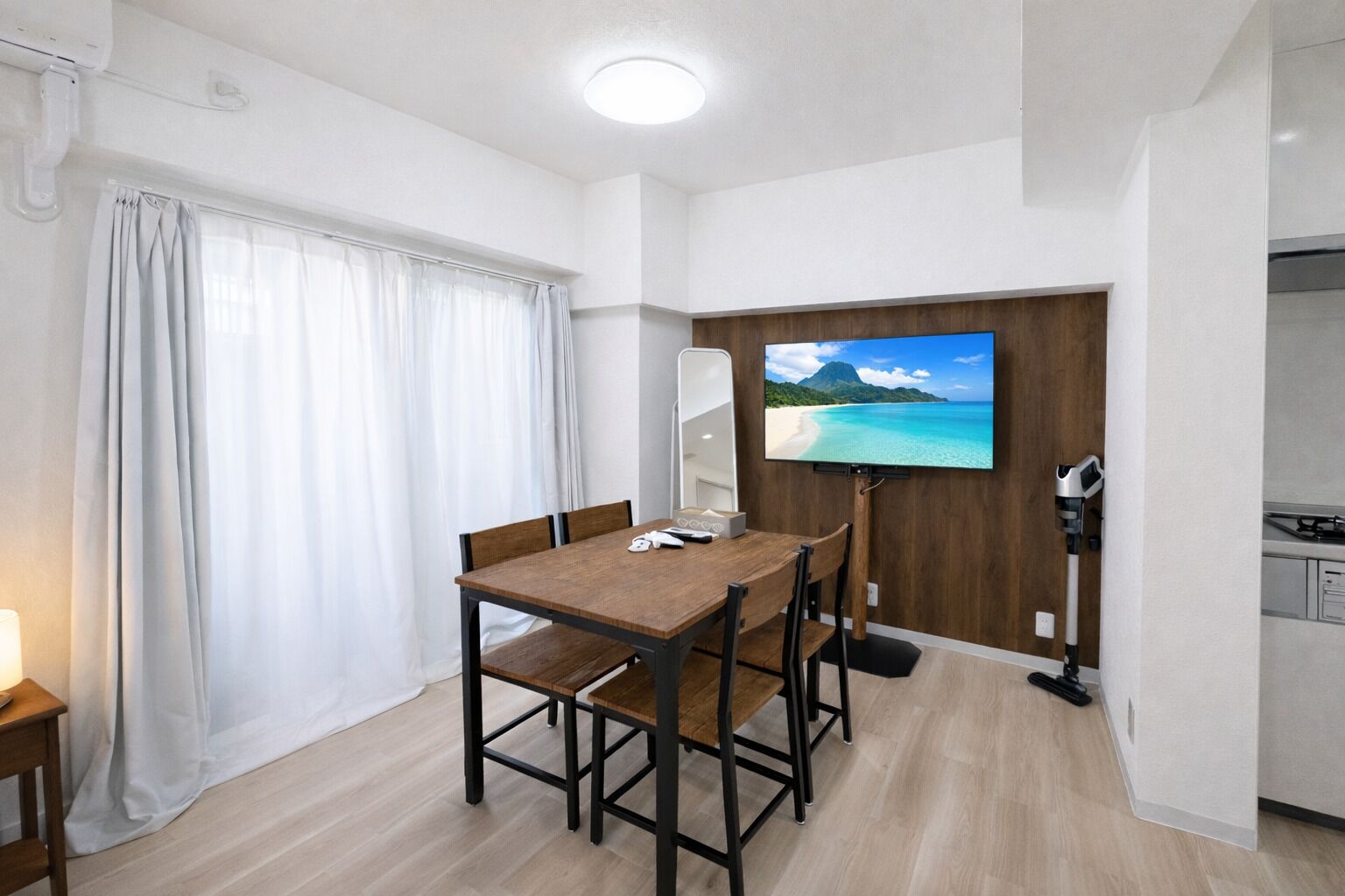 Una habitación compacta con una mesa de comedor de madera, un televisor de pantalla plana montado en un panel de madera, una pequeña kitchenette y un ventanal con cortinas.