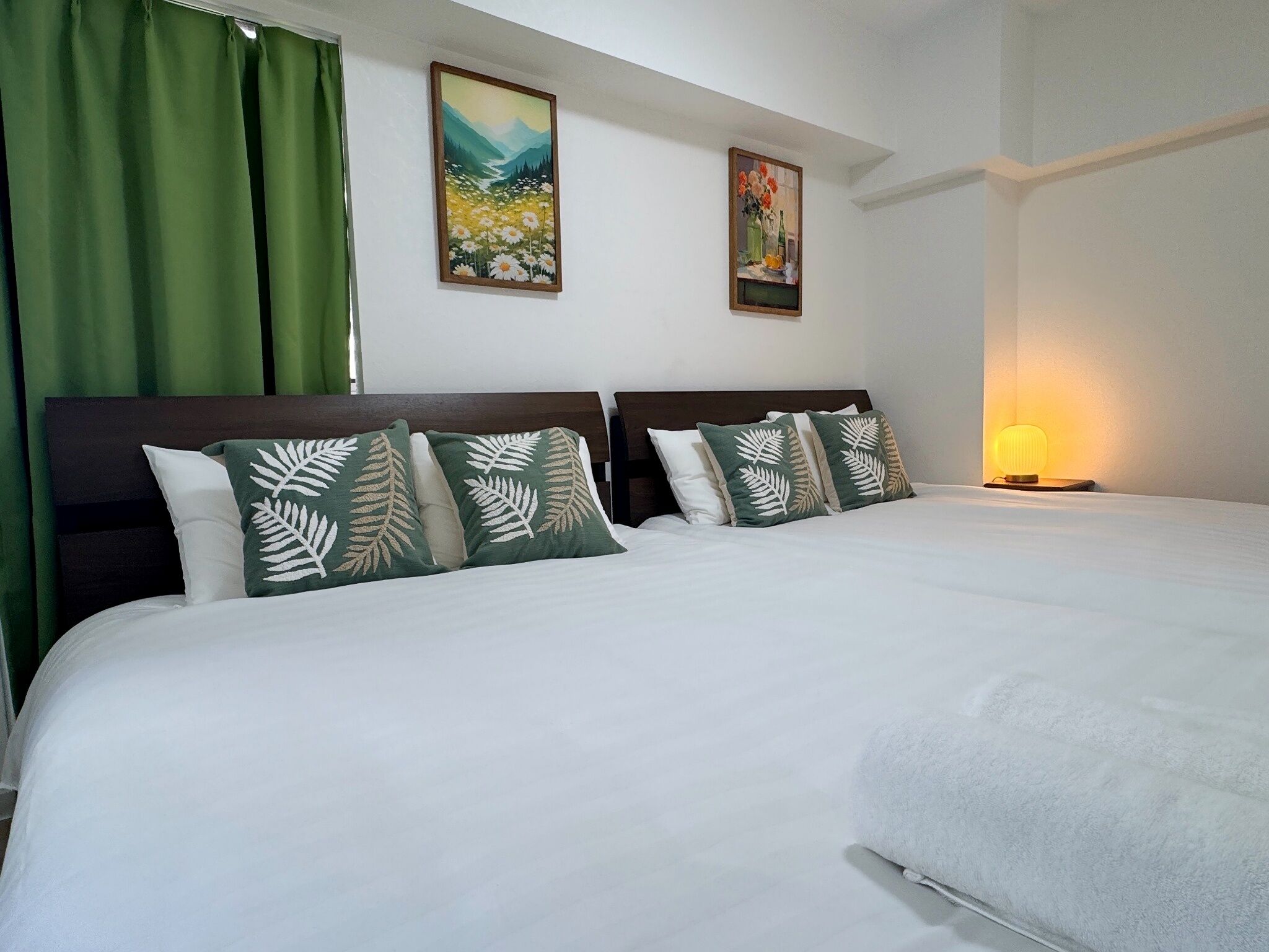 A hotel room with a large bed, green curtains, two paintings on the wall, and a bedside lamp.