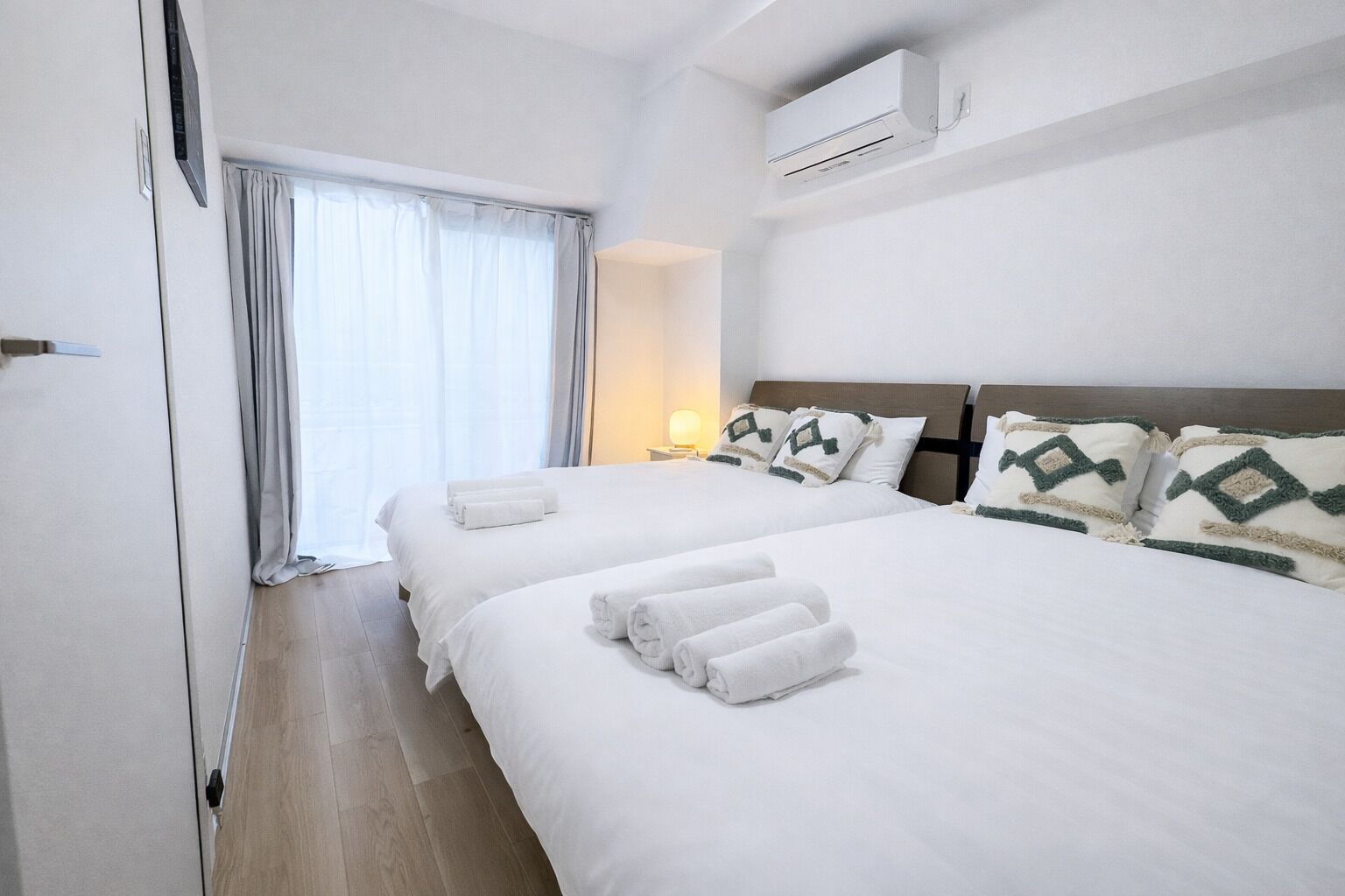 Habitación de hotel con dos camas, una mesita de noche con lámpara, aire acondicionado y una ventana con cortinas.