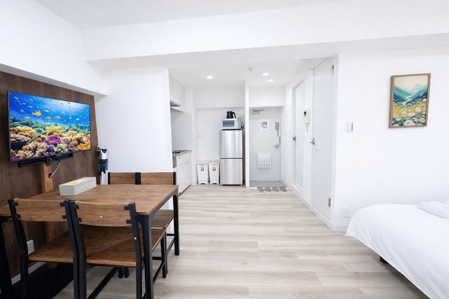 A modern, well-lit room with a wooden dining table, a flat-screen TV, a kitchen area with a refrigerator and microwave, and a bed with white bedding.