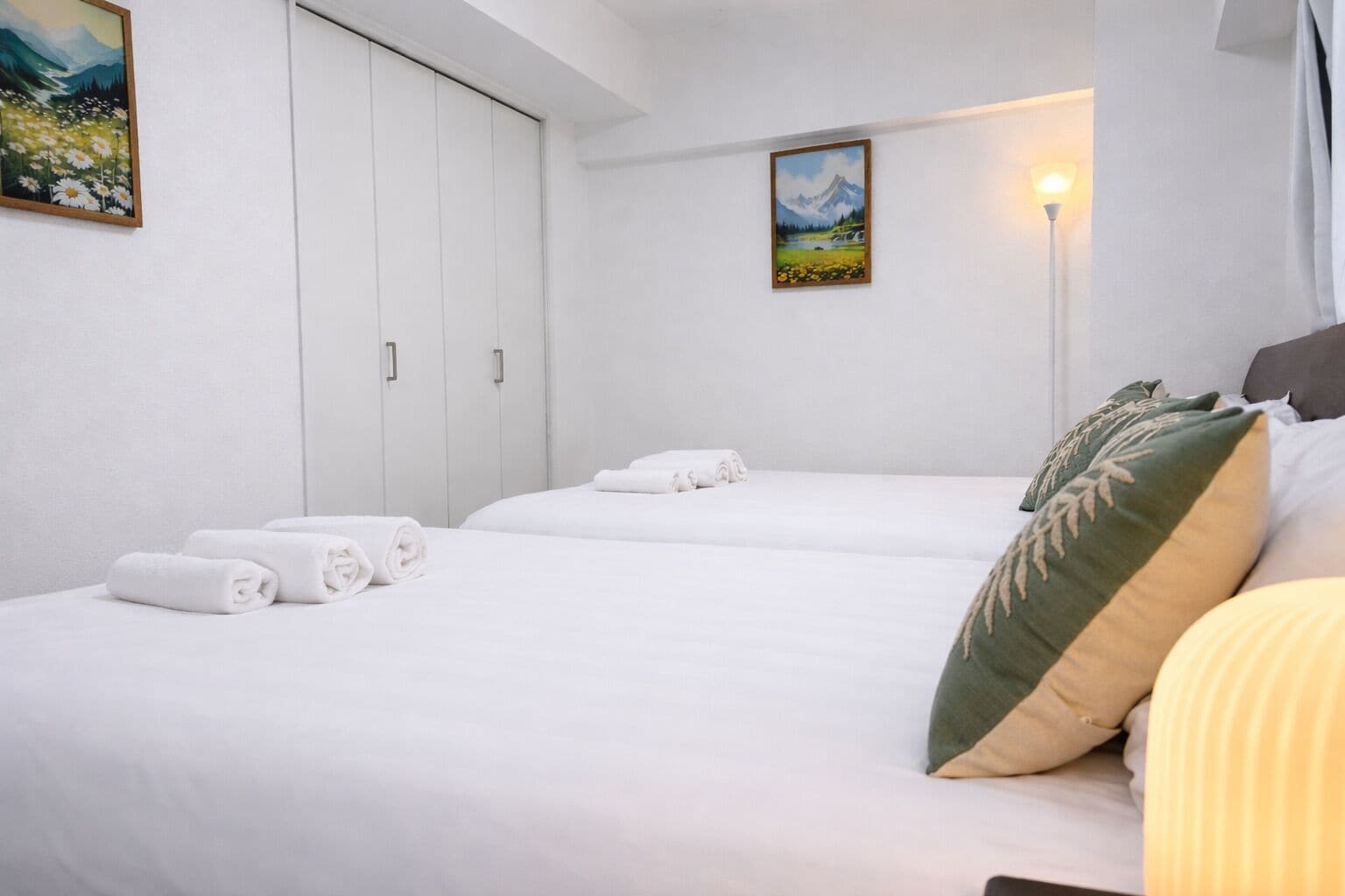 A hotel room with two beds, white bedding, a green pillow, a mountain painting on the wall, and a bedside lamp.