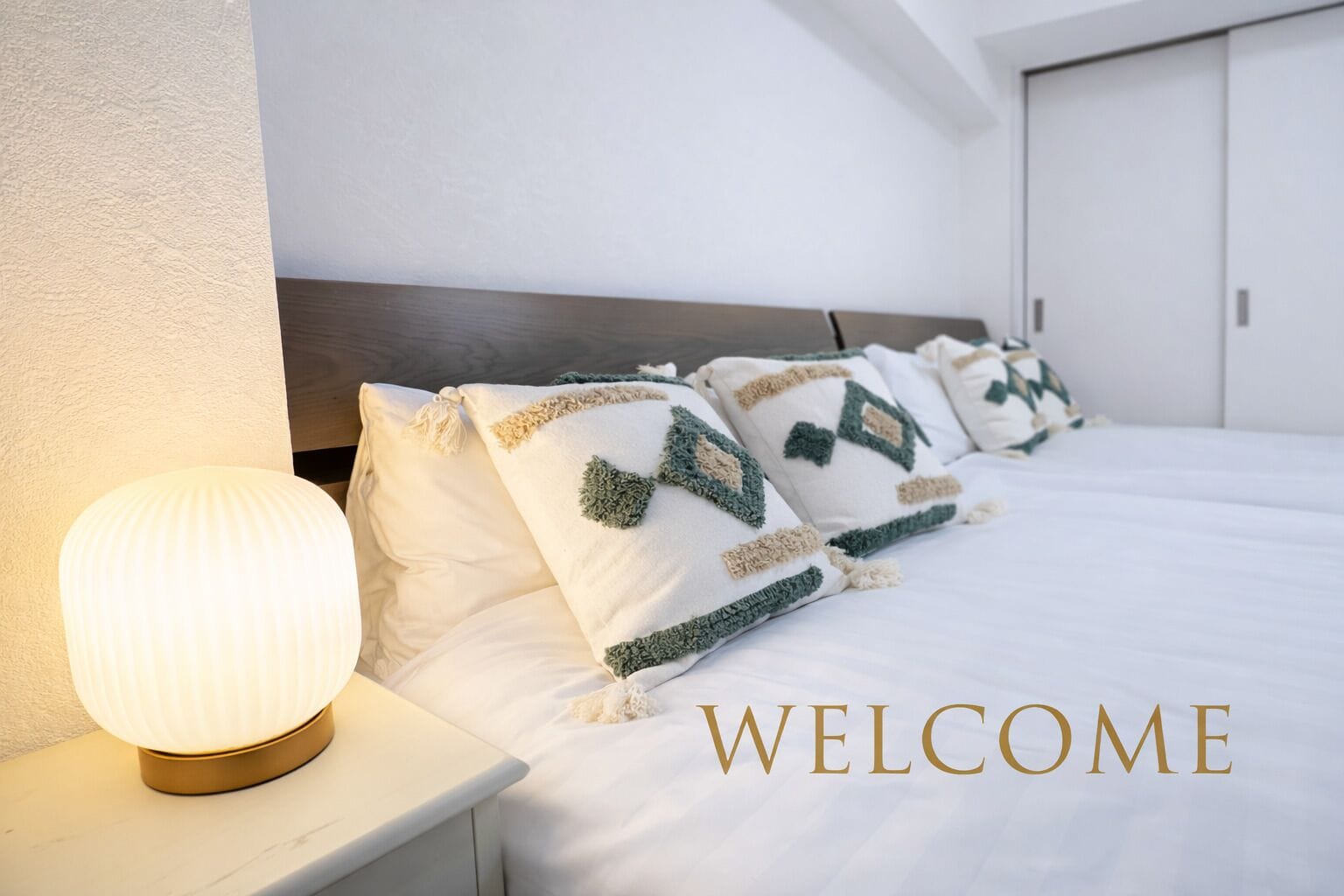 A neatly made bed with patterned pillows and a bedside lamp, with a "WELCOME" sign on the wall.