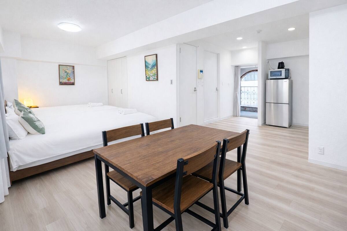 A modern, minimalist bedroom with a large bed, a wooden dining table, and a kitchenette in the background.