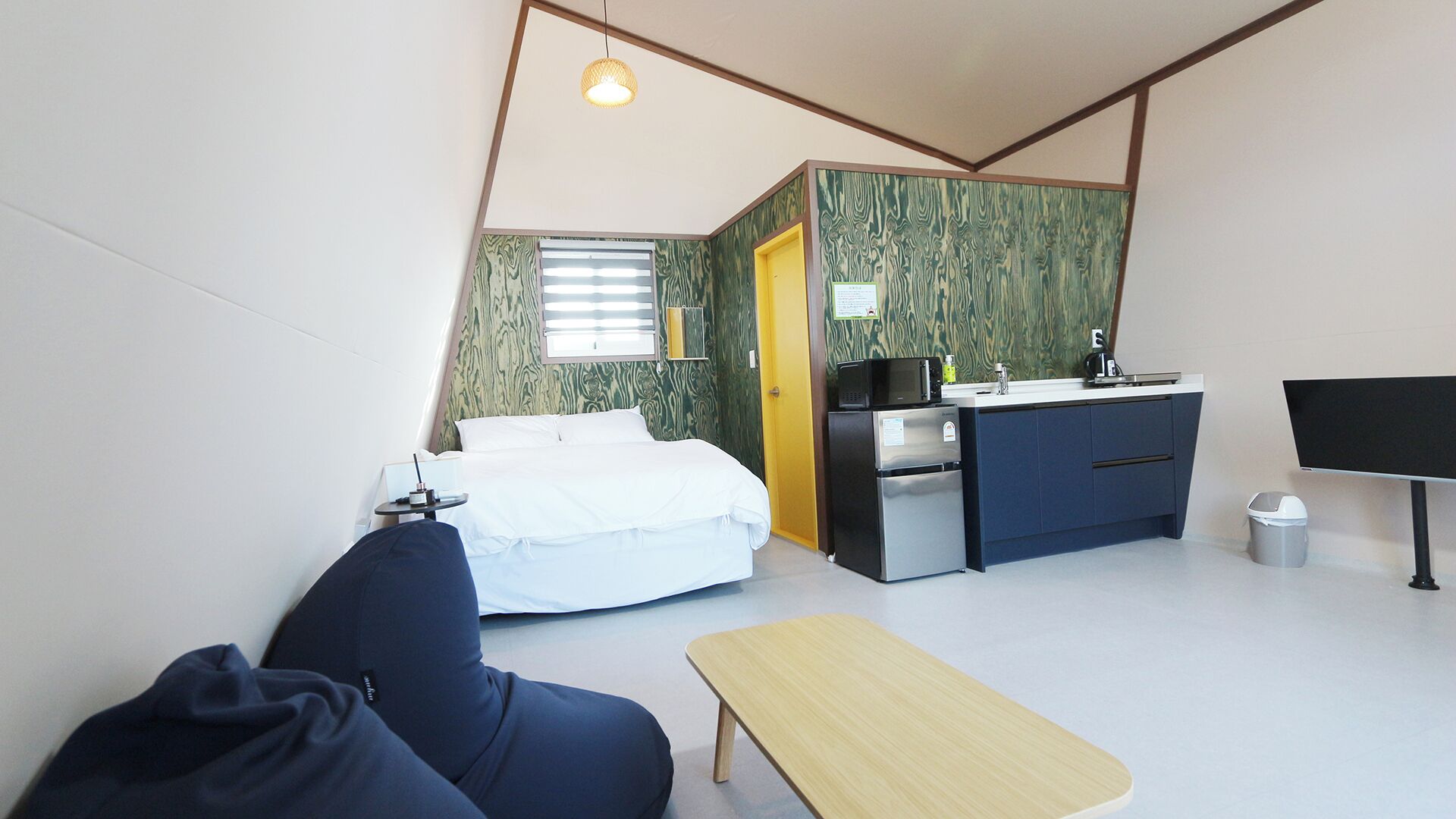 A compact hotel room with a bed, a small desk, a kitchenette, and a single chair.