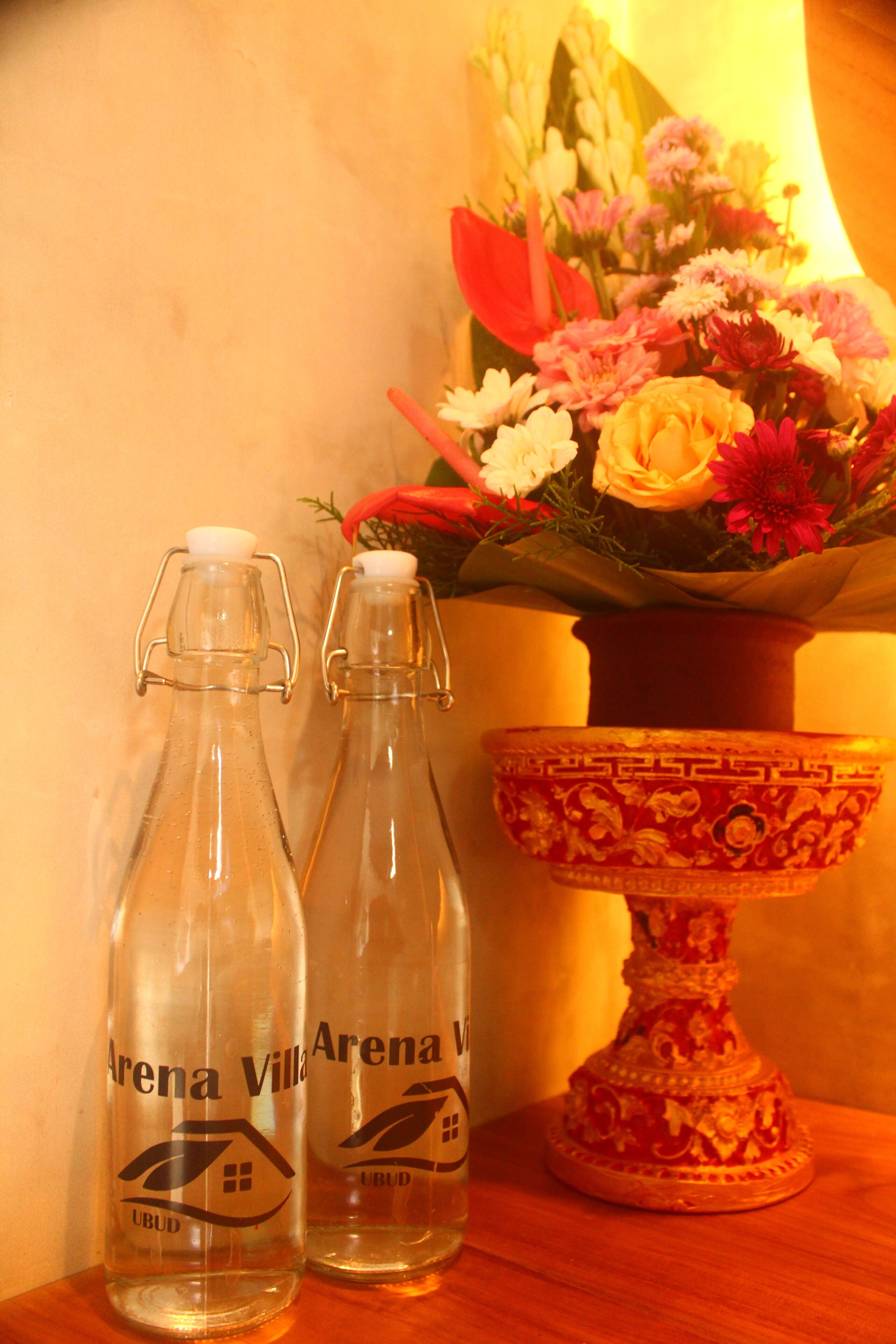 Dos botellas de vidrio con la etiqueta "Arena Villa", un cuenco decorativo y un arreglo floral al fondo.