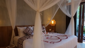 A bedroom with a canopy bed, a bedside table with a lamp, and a pair of slippers on the floor. - Arena Villa Ubud (Ubud)