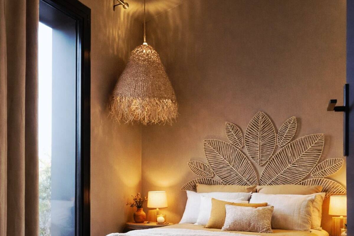 A bedroom with a large bed, a decorative leaf wall art, a hanging light fixture, and a view of the outdoors through a window.