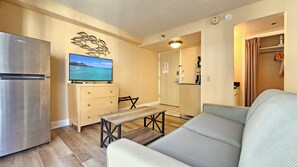 A compact living space with a kitchenette, a flat-screen TV mounted on a cabinet, a wooden table, a sofa, and a decorative wall piece. - Aloha Surf 706 — Central Waikiki Queen Studio-Suite w/Sofa-Bed&Kitchen ⁂Sleeps 3 (Honolulu)