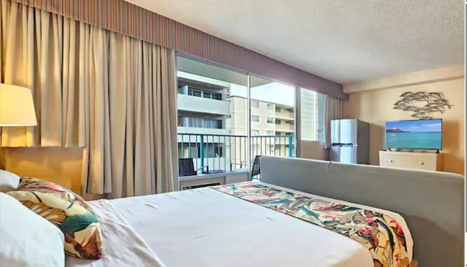 A hotel room with a large bed, a desk, a chair, a TV, and a window with a view of a building.