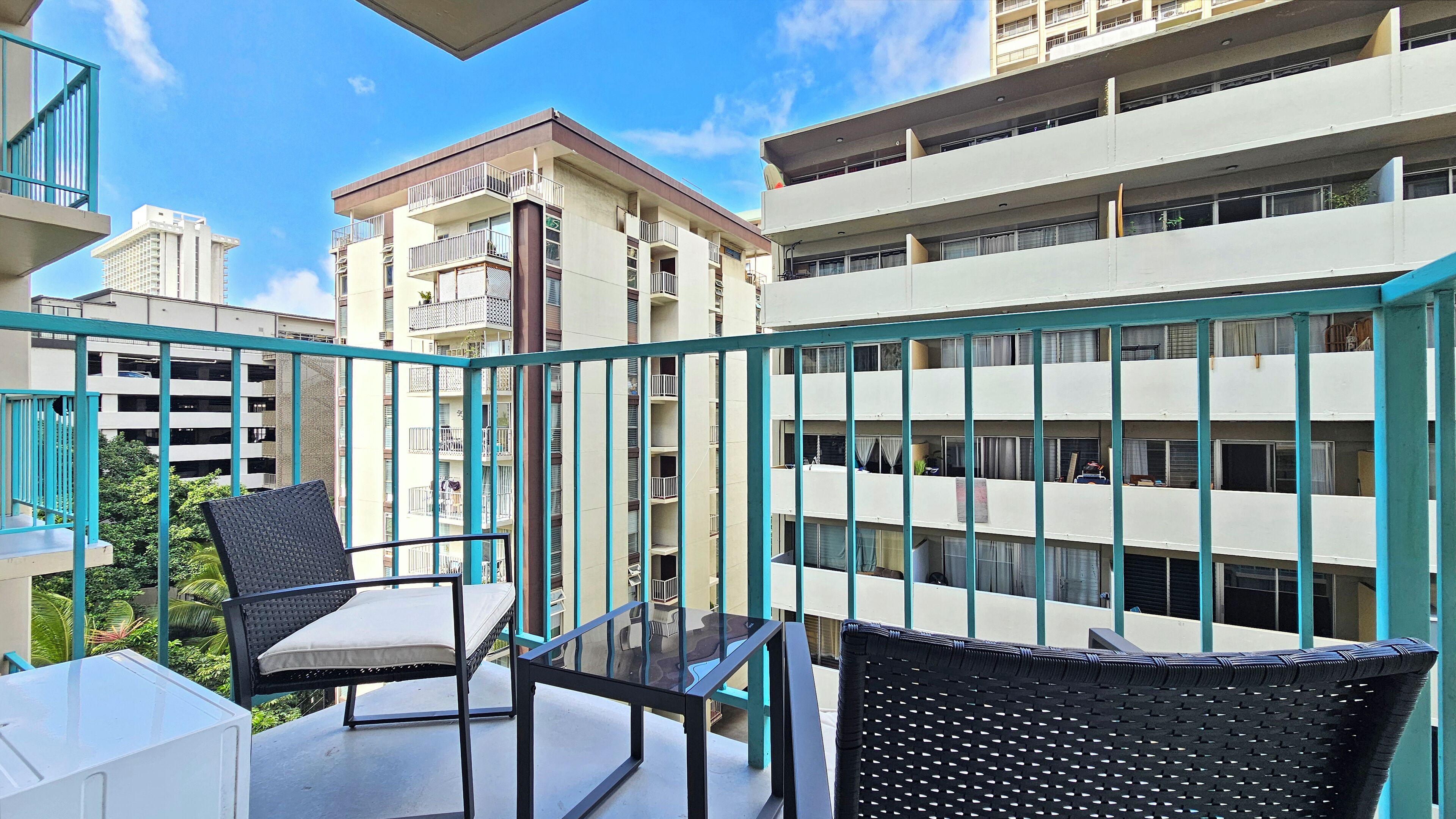 Ein Balkon mit zwei Korbstühlen und einem kleinen Tisch, der auf ein Gebäude mit mehreren Balkonen und einem strahlend blauen Himmel blickt.