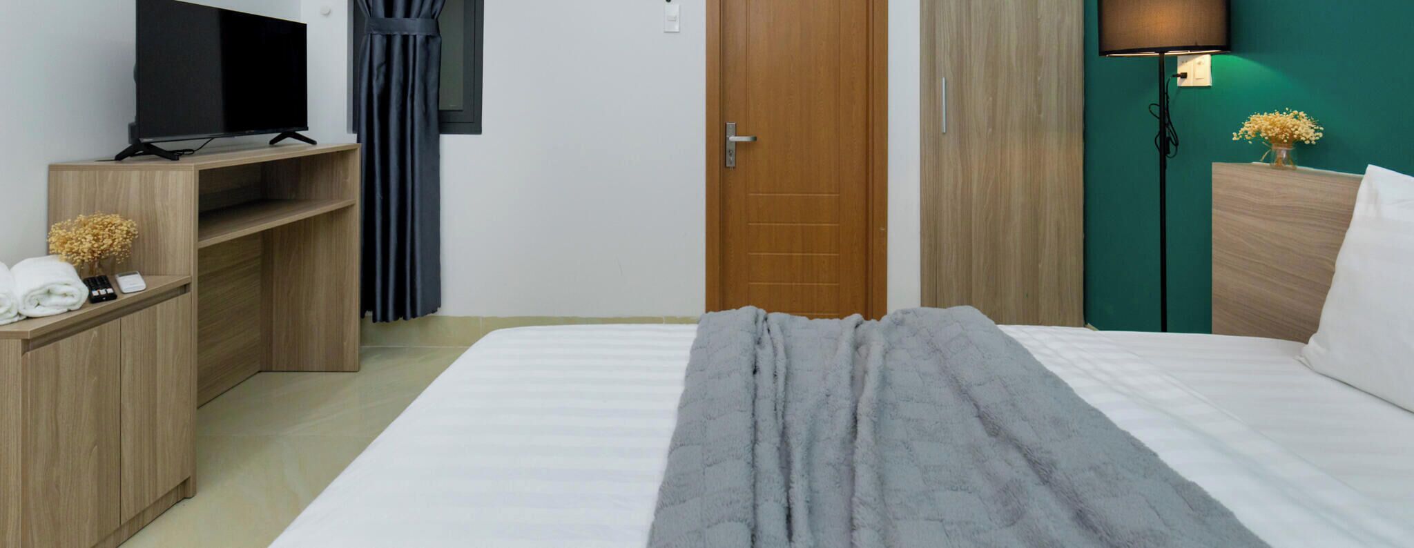 A hotel room with a bed, a TV on a wooden stand, a wooden wardrobe, a door, and a nightstand with towels.