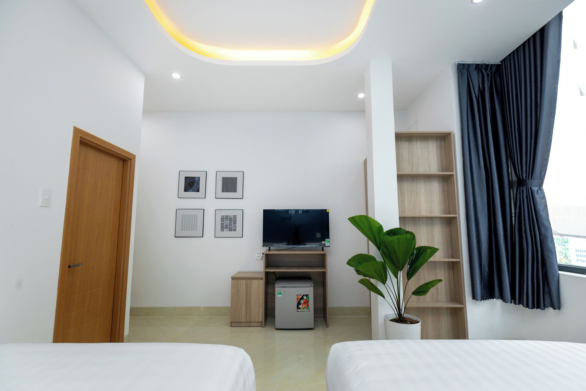 Habitació d'hotel amb un llit, una taula de treball amb un ordinador, una nevera petita, un televisor, una planta i una finestra amb cortines.