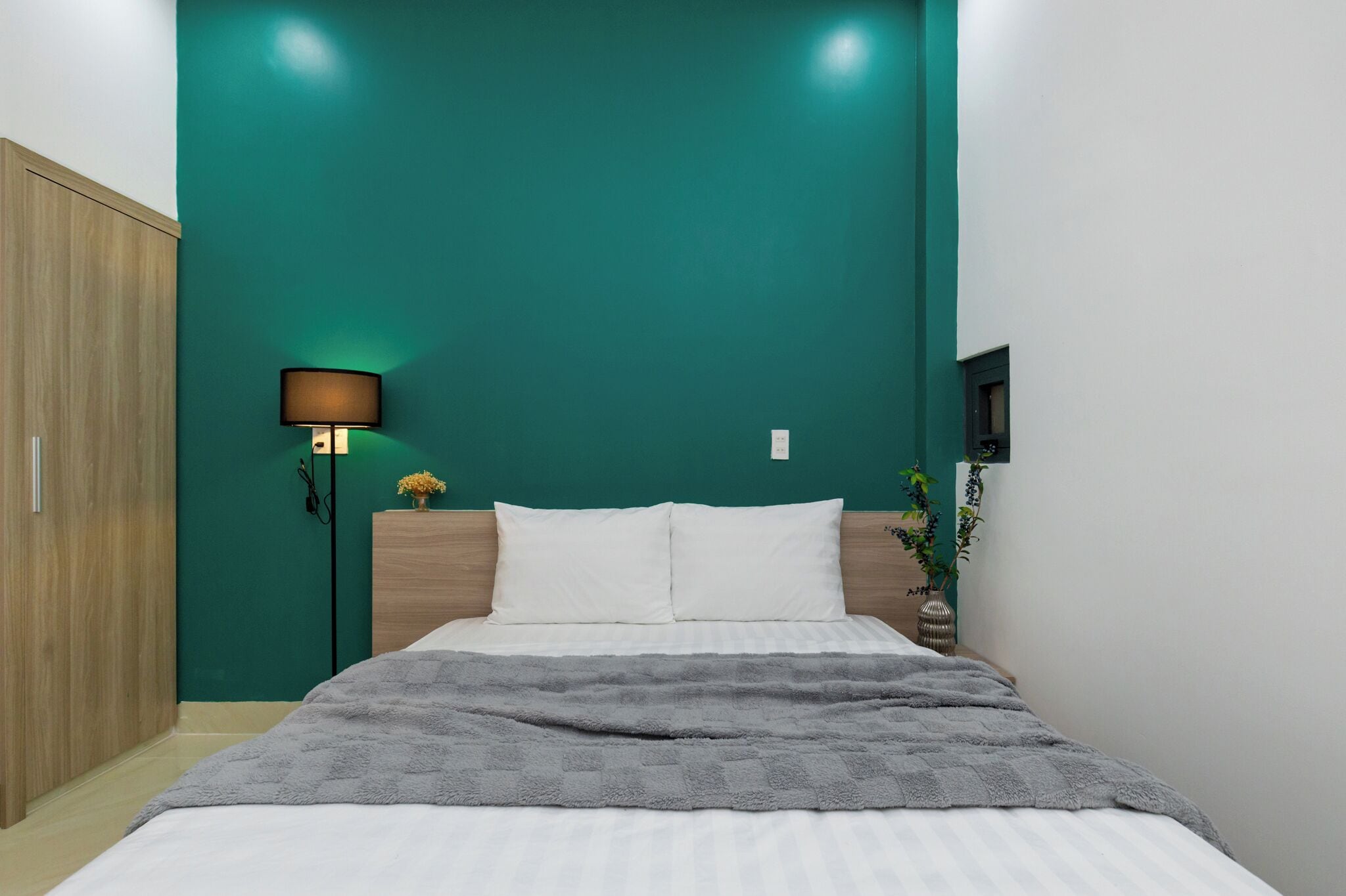 A bedroom with a teal accent wall, a wooden wardrobe, a bedside lamp, and a bed with white bedding.