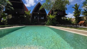 Ein klares Schwimmbecken, umgeben von traditionellen Holzhäusern mit roten Dächern, Palmen und einem strahlend blauen Himmel.