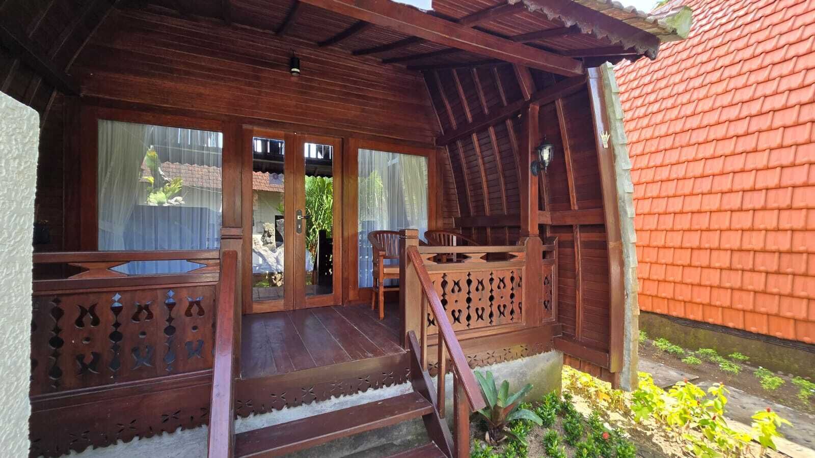 Un porche de madera con un banco, un pequeño jardín y un edificio de techo rojo al fondo.