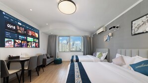 Een moderne hotelkamer met een grote tv waarop verschillende zenders te zien zijn, een zithoek met een tafel en stoelen, een bank en een groot raam met uitzicht over de stad.