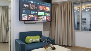 Een moderne woonkamer met een flatscreen-televisie aan de muur, waarop diverse programma’s te zien zijn. Een blauwe bank met een groen kussen en een ‘cool’ kussen staat voor de televisie. Een kleine tafel met een bloevas en een fles is voor de bank geplaatst. De kamer heeft een raam met gordijnen en uitzicht op het buiten.