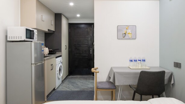 Habitación de hotel compacta con cama, una pequeña mesa con silla, microondas, refrigerador, lavadora y puerta que da a un pasillo.