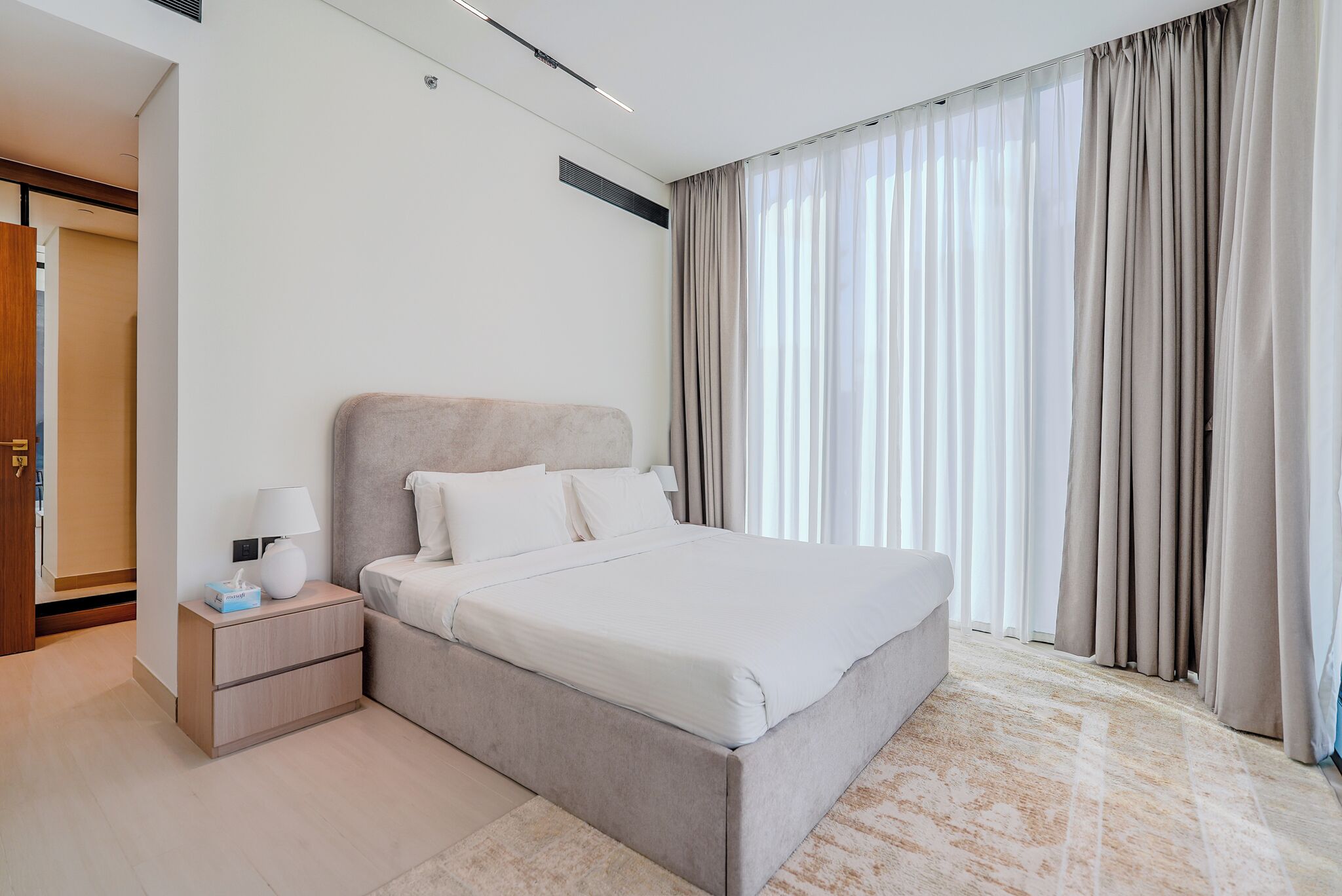 A modern hotel room with a large bed, a bedside table with a lamp, and a view of the city through sheer curtains.