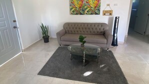 Un salon avec un canapé beige, une table basse en verre ornée d’une plante, un tableau abstrait aux couleurs vives et un panneau indiquant le mot de passe du réseau Wi-Fi, accroché au mur.