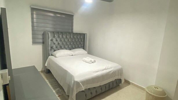 Une chambre à coucher comprenant un lit, un ventilateur de plafond, une fenêtre avec des stores, une table de chevet et une poubelle.