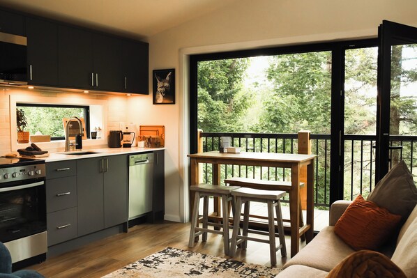 A modern kitchen with a dining area, a large window, and a view of greenery. - Tinwood Cabins The Descent, Cozy 3 min to Rossland (Rossland)